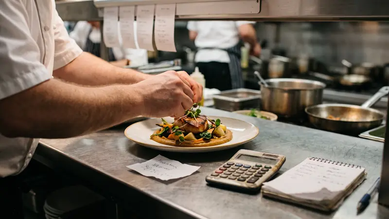 Chef finalizando prato ao lado de calculadora e bloco de anotações para definir o preço