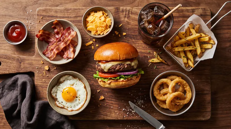 Hambúrguer com adicionais de bacon, cheddar, ovo e anéis de cebola em tigelas ao redor